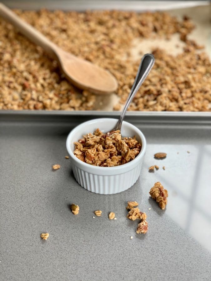 Small white ramekin of Crunchy Tahini Granola with silver spoon in front of a tray of granola.