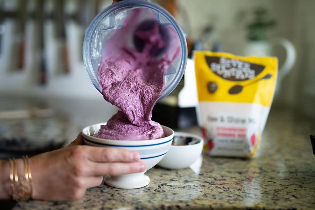 Pouring thick, purple berry smoothie in a white bowl on marble countertop.