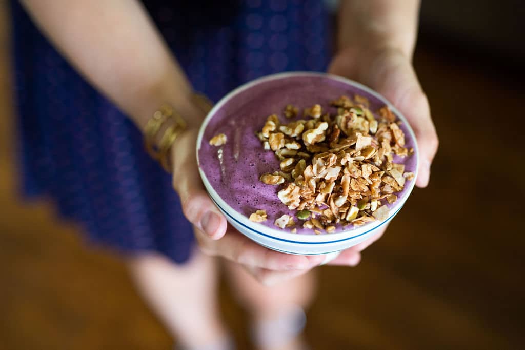 Two hands holding a bowl with purple berry smoothie and granola topping.