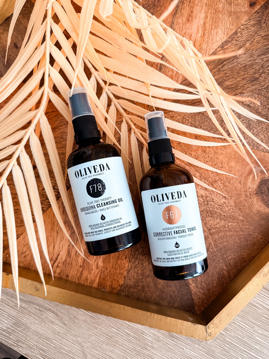 Oliveda F78 Arbequina Cleansing Oil and F67 Corrective Facial Tonic on a wooden tray with a palm leaf