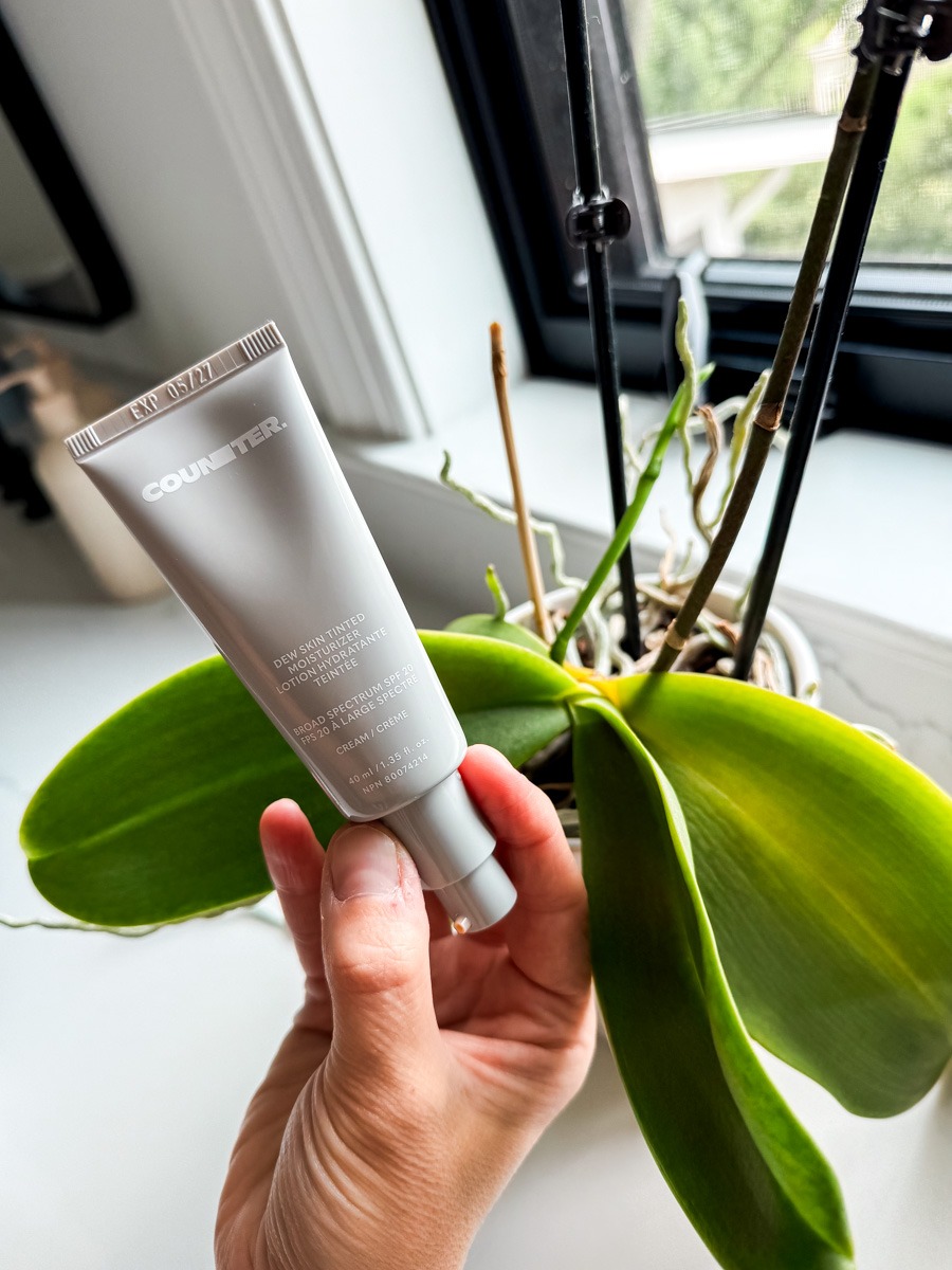 Counter Dew Skin Tinted Moisturizer in hand with an orchid in back