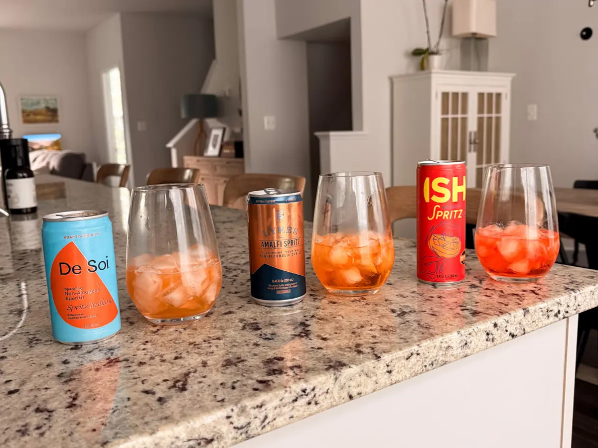 uma fileira de spritzes de aperol sem álcool: de sio, Lyre's, Ish, todos servidos em taças de vinho com gelo na cozinha de Kath